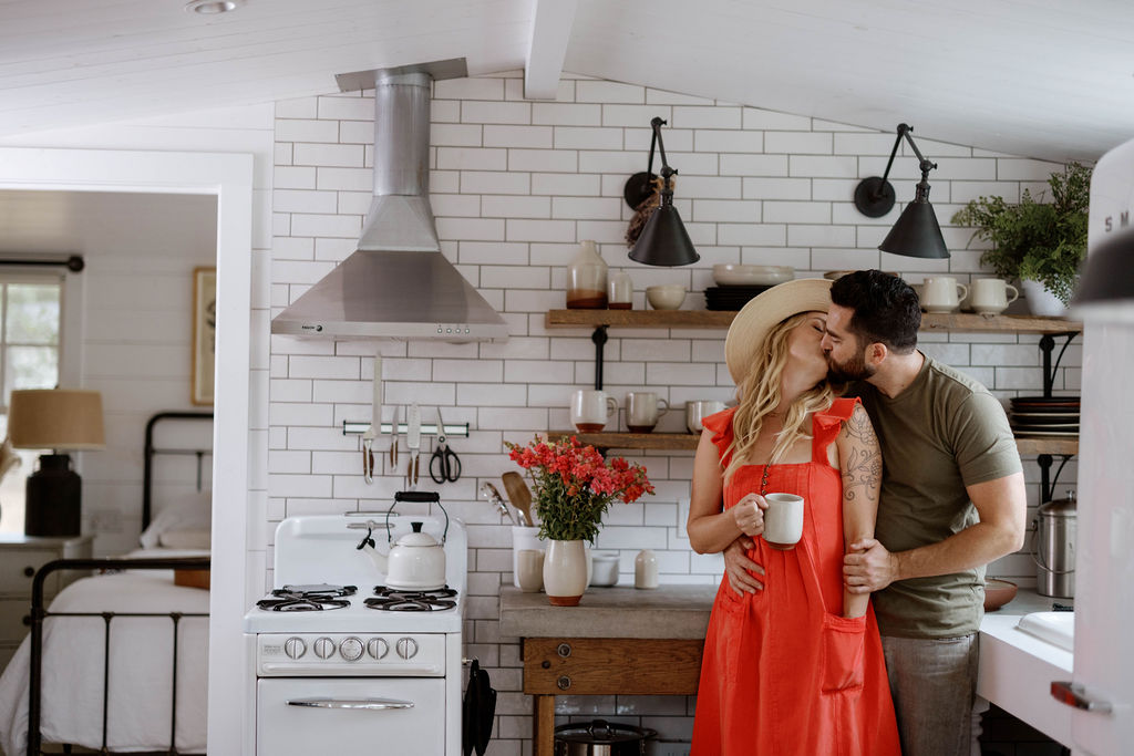 A couple sharing a kiss over morning coffee in a sunlit kitchen filled with handmade ceramics